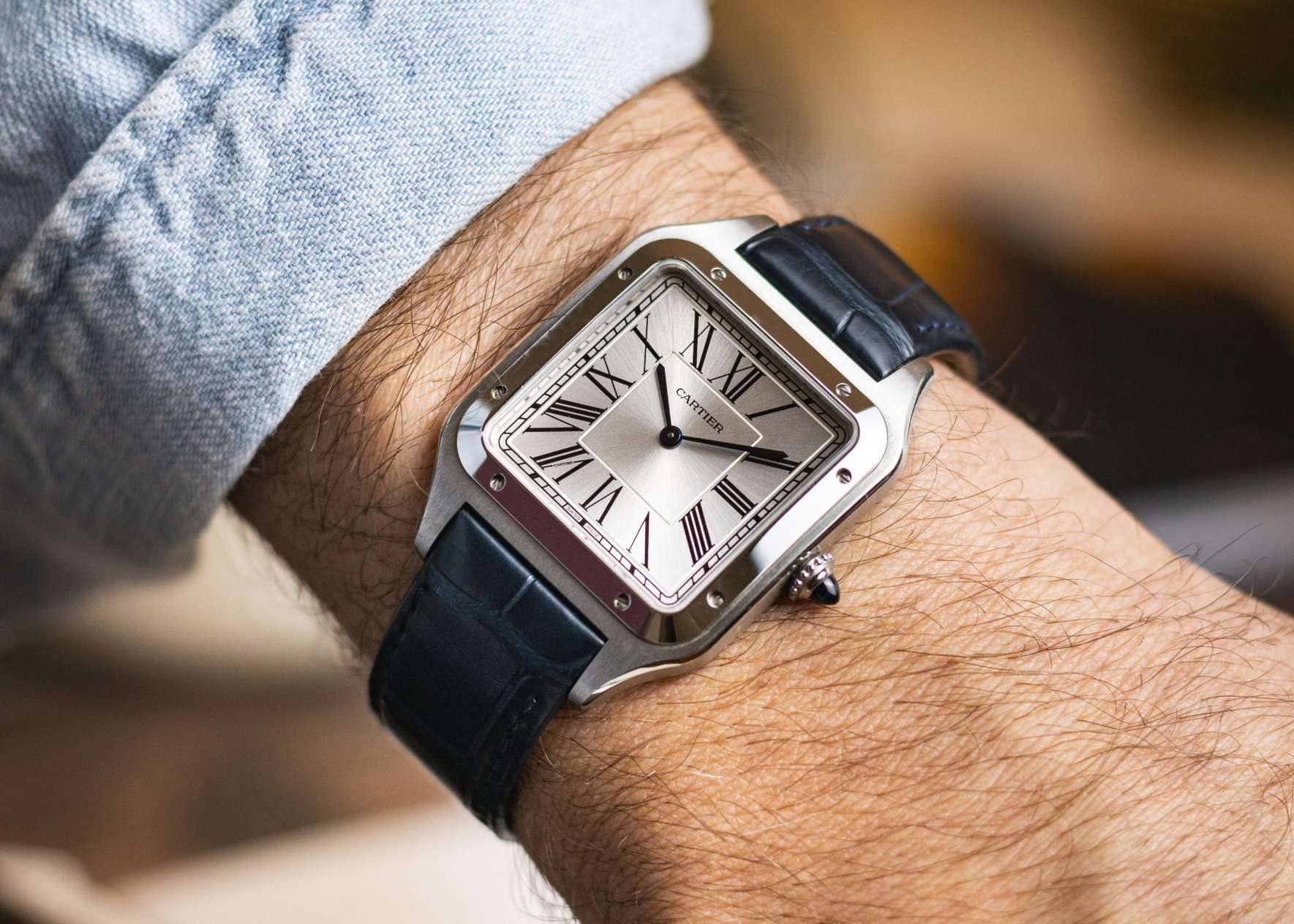 Cartier Santos-Dumont mit silbernem Zifferblatt und blauem Lederarmband abgebildet an einem Handgelenk mit hellblauem Ärmel vor braunem Hintergrund