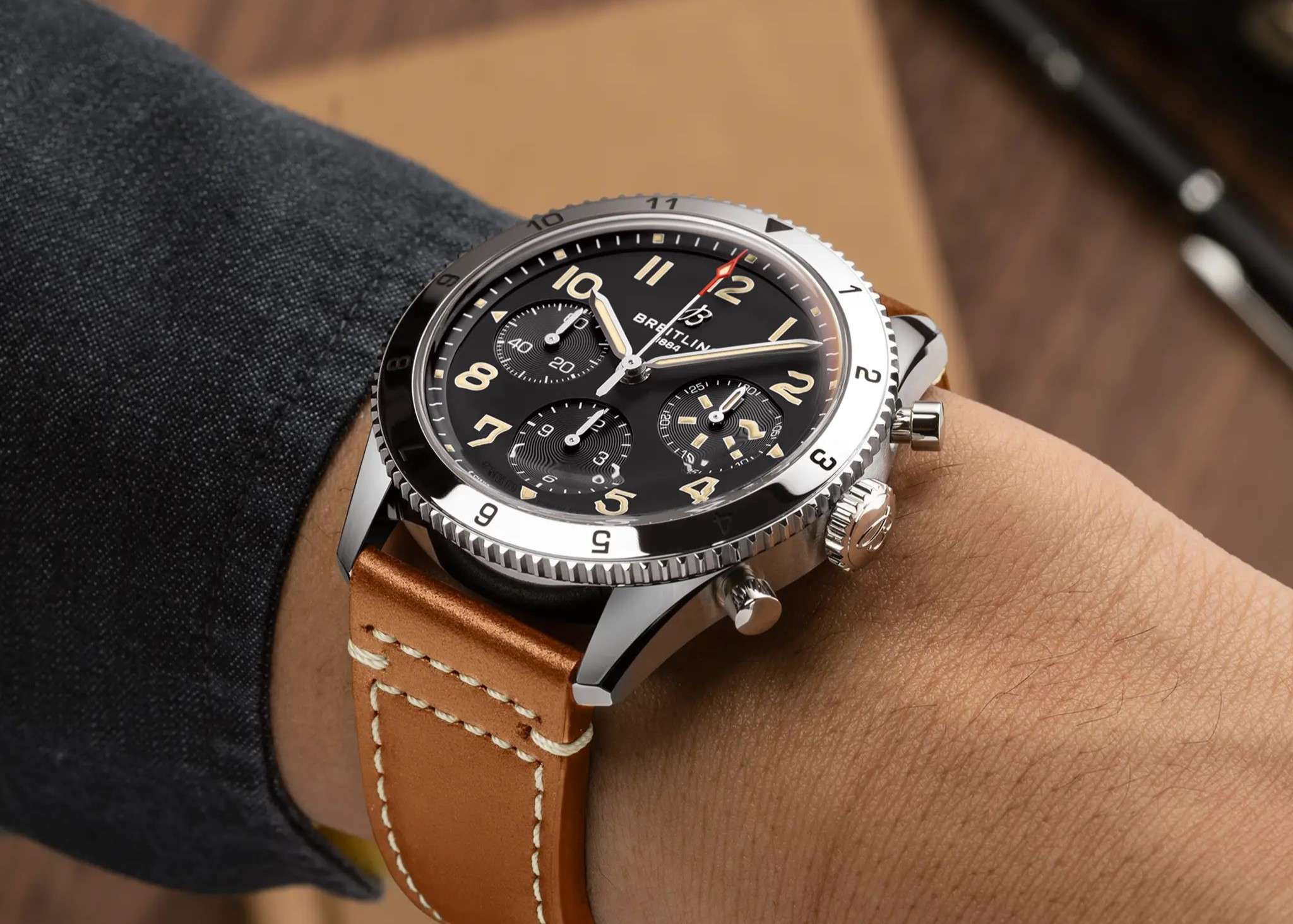 Breitling Classic Avi Chronograph 42 P-51 Mustang mit schwarzem Zifferblatt und braunem Lederarmband abgebildet an einem Handgelenk mit schwarzem Ärmel vor beige-braunem Hintergrund