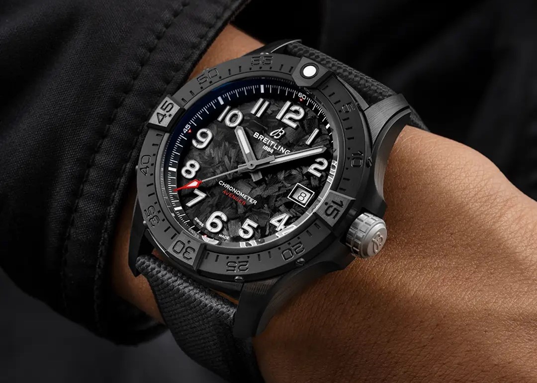 Breitling Avenger Automatic 42 Night Mission mit schwarzem Zifferblatt, schwarzem Gehäuse und schwarzem Lederarmband abgebildet an einem Handgelenk mit schwarzem Ärmel vor schwarzem Hintergrund