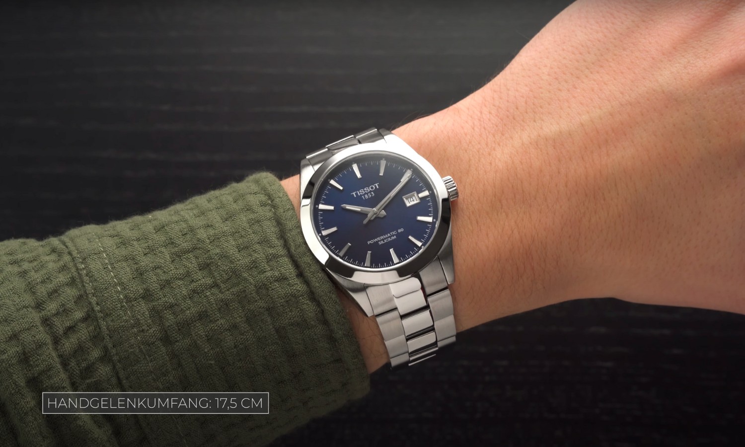 Die Tissot Gentleman Powermatic 80 mit blauem Zifferblatt am Edelstahlarmband an einem Handgelenk mit grünem Ärmel getragen vor schwarzem Hintergrund