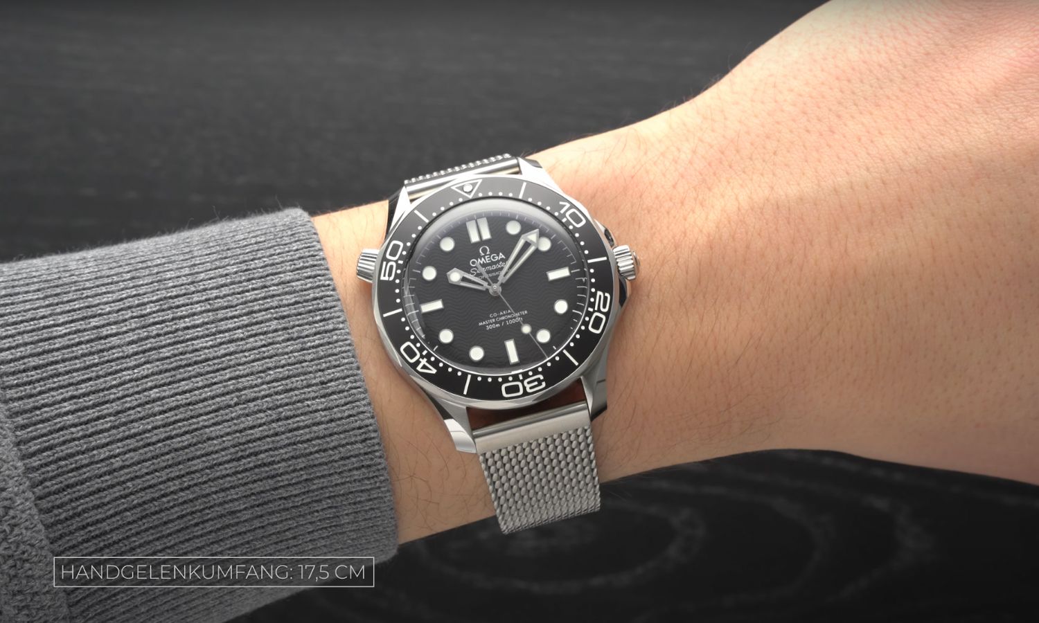 Die OMEGA Seamaster Diver 300M No-Date mit schwarzem Zifferblatt am Milanaise-Armband an einem Handgelenk mit grauen Ärmel getragen vor dunkelgrauem Hintergrund