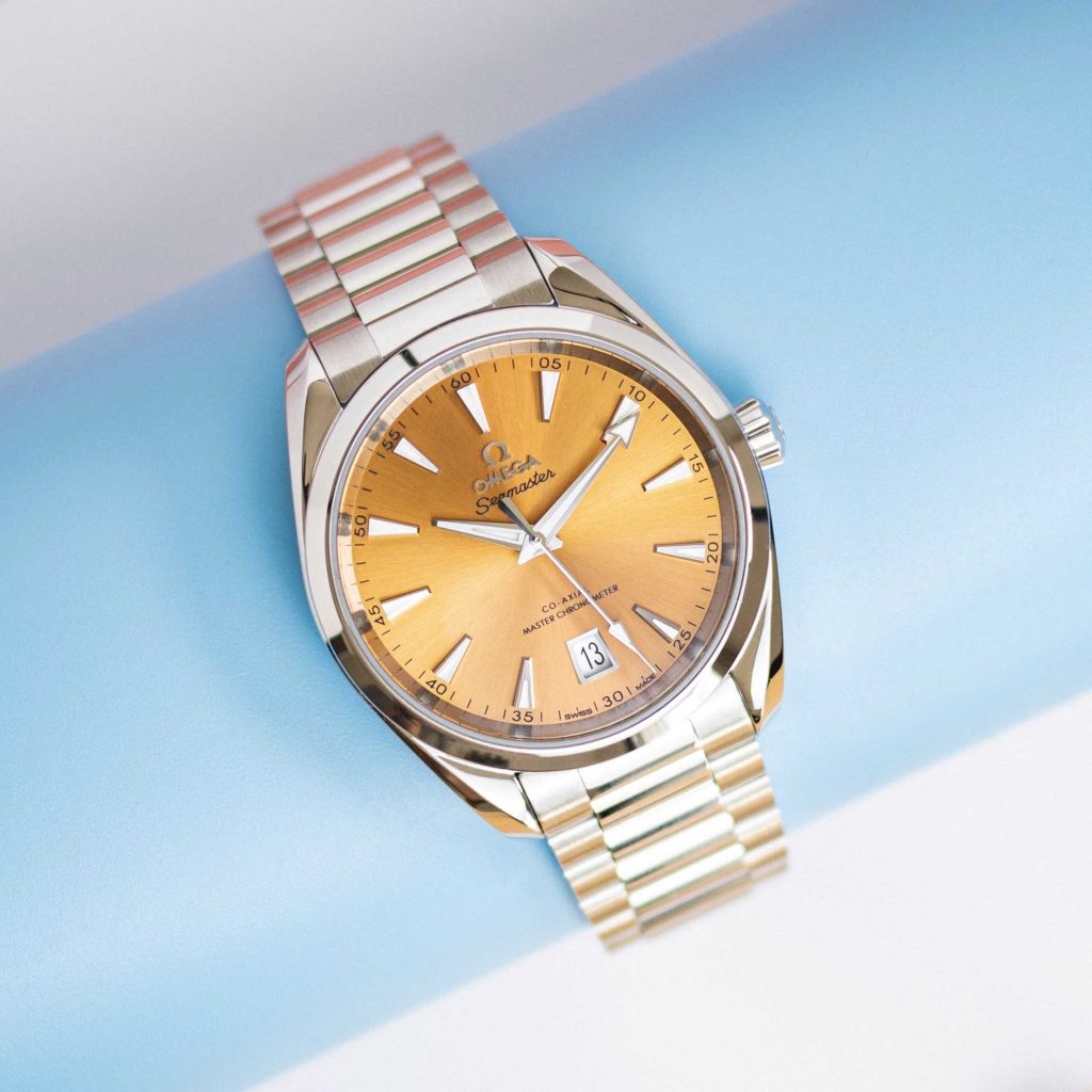 Omega Seamaster Aqua Terra mit gelbem Zifferblatt am Edelstahlarmband abgebildet vor weißblauem Hintergrund