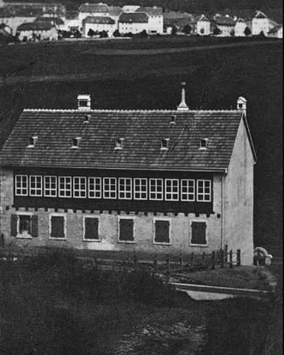 Schwarzweiß-Foto der Fabrik von Longines, die auf einem Feld gelegen ist, von dem aus die Häuser im Hintergrund zu sehen sind