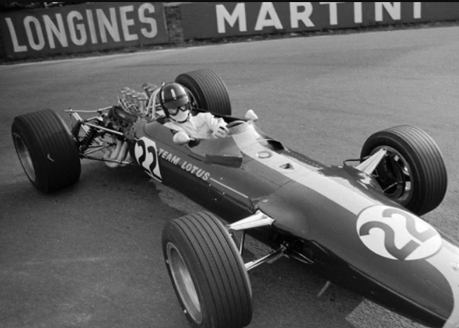 Schwarzweiß-Foto eines alten Formel1-Wagens und einem Fahrer, der darin sitzt. An der Bande im Hintergrund prangt der Longines-Schriftzug