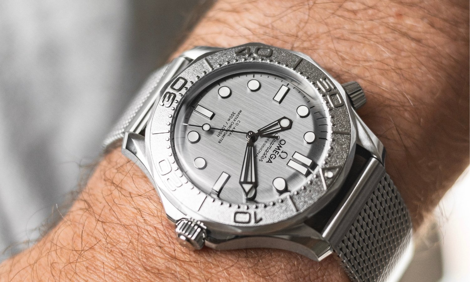 OMEGA Seamaster Diver 300M Monochrome mit grauem Zifferblatt am Milanaise-Armband an einem Handgelenk mit grauem Ärmel getragen vor grauem Hintergrund