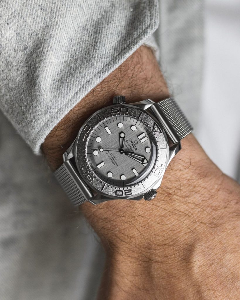 OMEGA Seamaster Diver 300M Monochrome mit grauem Zifferblatt am Milanaise-Armband an einem Handgelenk mit grauem Ärmel getragen vor grauem Hintergrund
