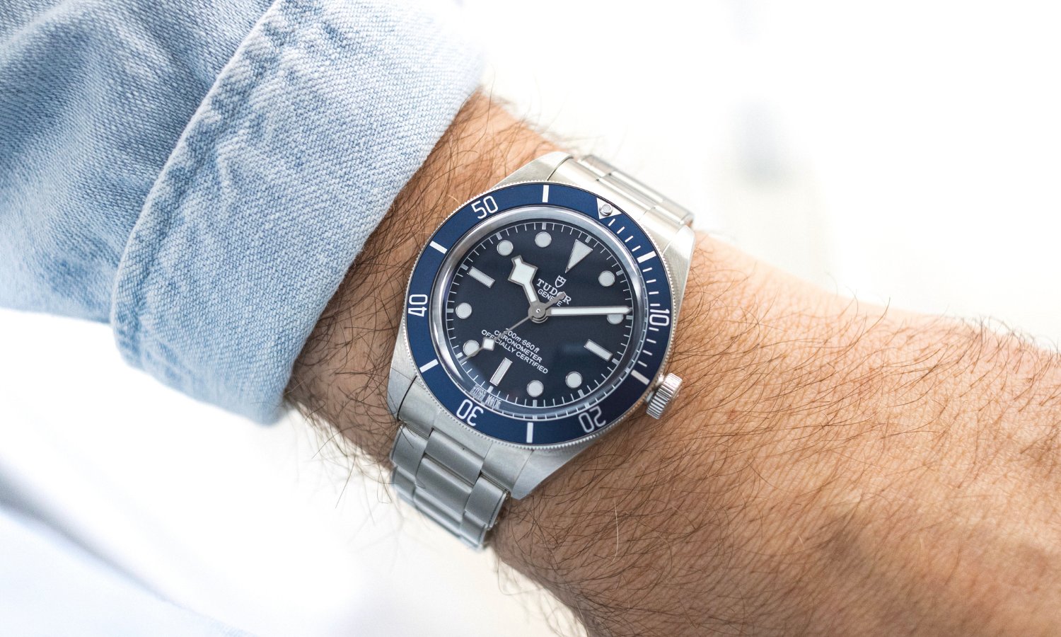 TUDOR Black Bay 58 mit blauem Zifferblatt am Edelstahlarmband an einem Handgelenk mit hellblauem Hemdärmel getragen vor hellem Hintergrund