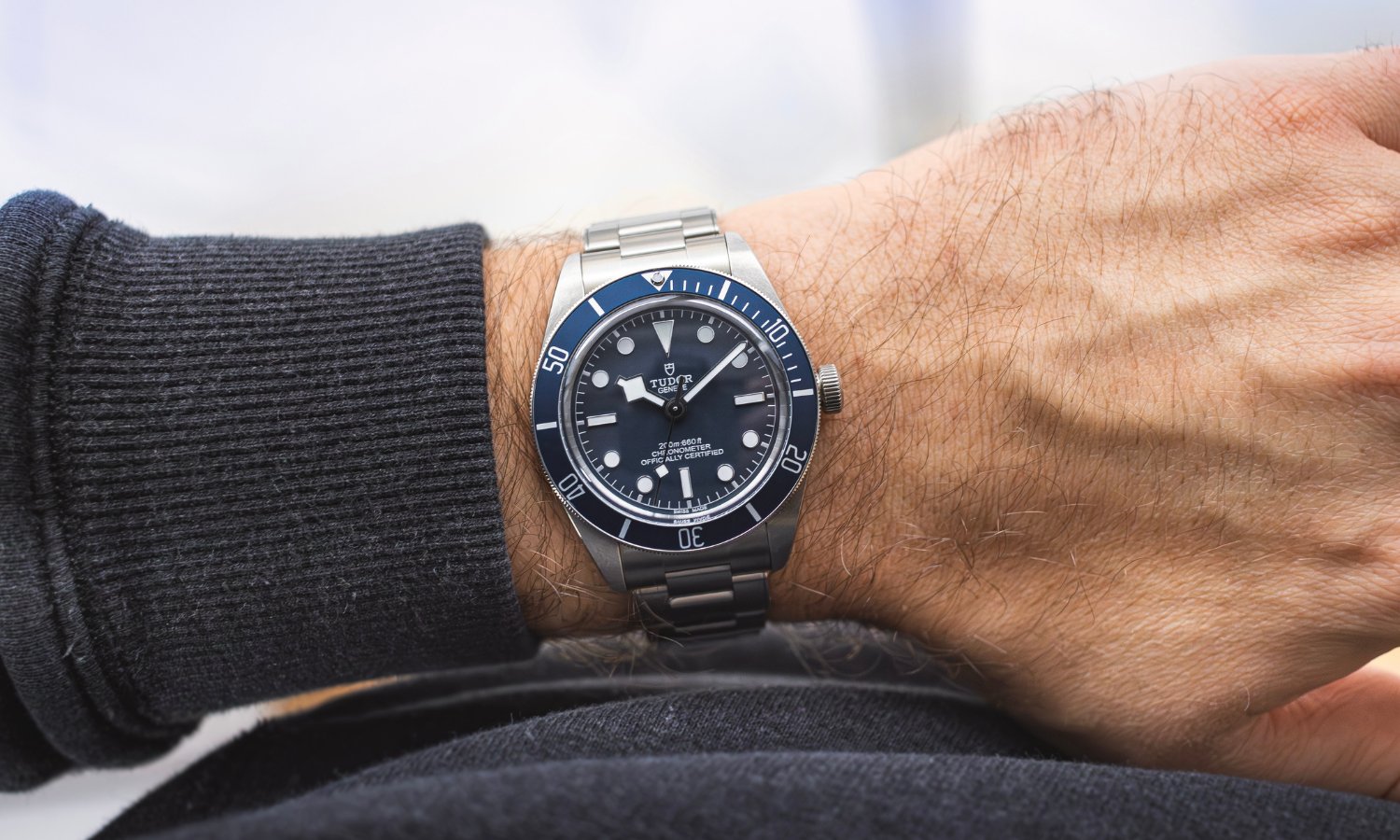 TUDOR Black Bay 58 mit blauem Zifferblatt am Edelstahlarmband an einem Handgelenk mit dunkelgrauem Ärmel getragen vor hellem Hintergrund