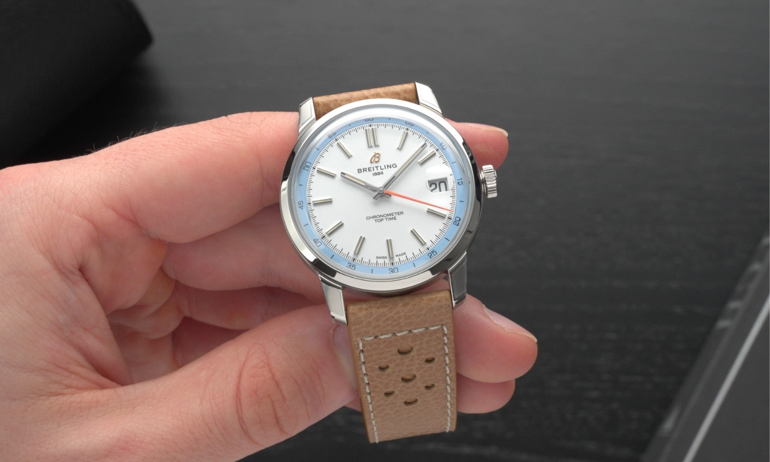 Die Breitling Top Time B31 mit weißem Zifferblatt am Lederarmband in einer Hand gehalten vor dunkelgrauem Hintergrund