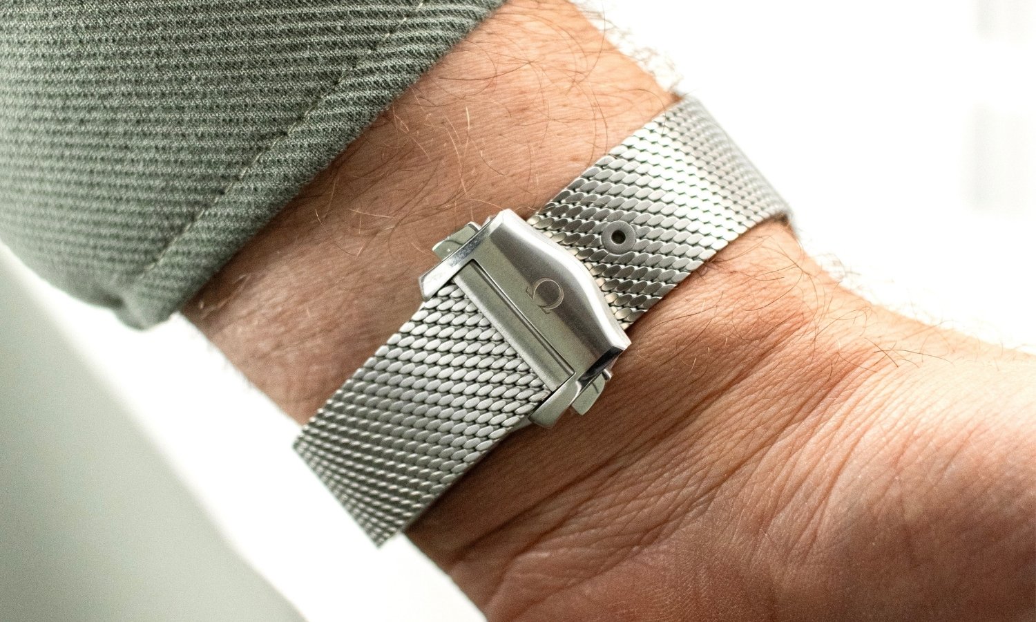 Das Milanaise-Edelstahlarmband der OMEGA Seamaster Diver 300M No-Date an einem Handgelenk mit grünem Ärmel getragen vor hellem Hintergrund