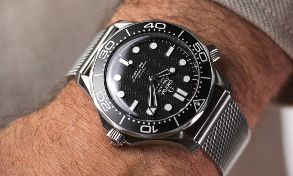 OMEGA Seamaster Diver 300M No-Date mit schwarzem Zifferblatt am Milanaise-Armband an einem Handgelenk mit grünem Ärmel getragen vor hellem Hintergrund