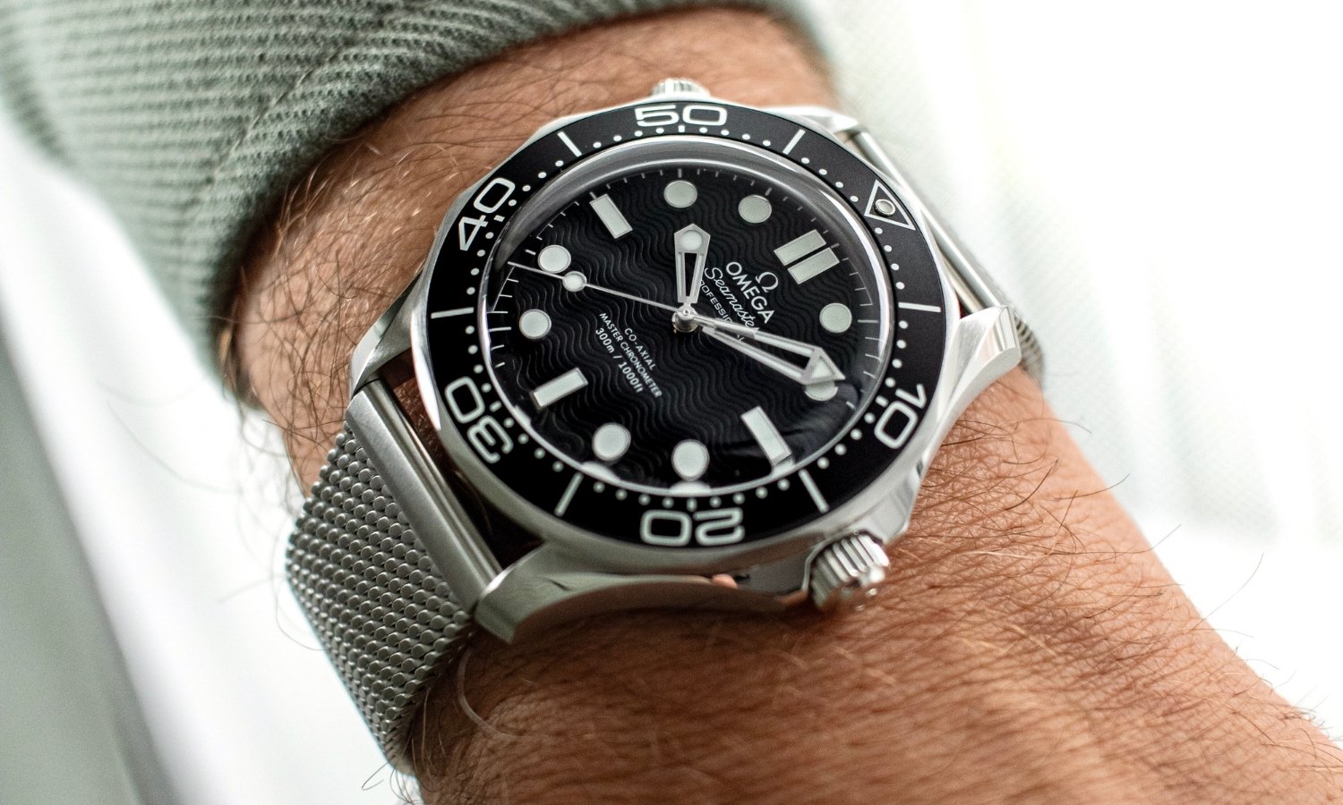 OMEGA Seamaster Diver 300M No-Date mit schwarzem Zifferblatt am Milanaise-Armband an einem Handgelenk mit grünem Ärmel getragen vor hellem Hintergrund