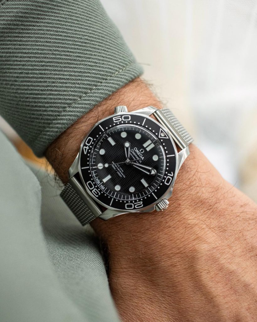 OMEGA Seamaster Diver 300M No-Date mit schwarzem Zifferblatt am Milanaise-Armband an einem Handgelenk mit grünem Ärmel getragen vor hellem Hintergrund