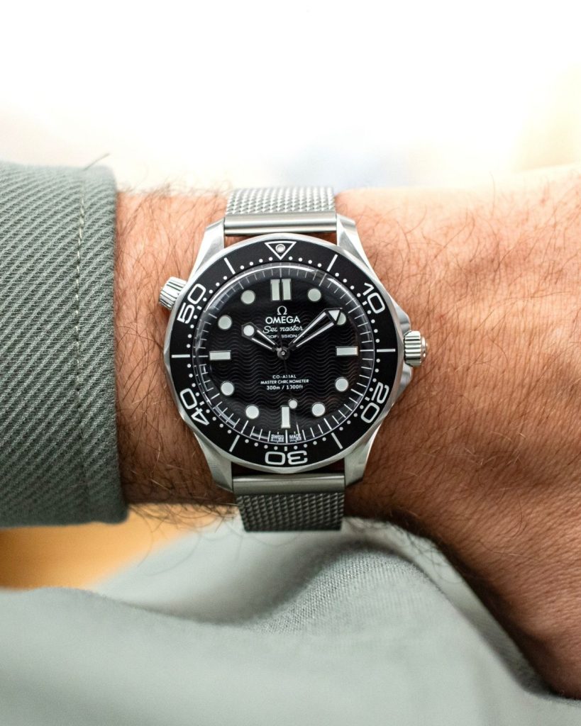 OMEGA Seamaster Diver 300M No-Date mit schwarzem Zifferblatt am Milanaise-Armband an einem Handgelenk mit grünem Ärmel getragen vor hellem Hintergrund