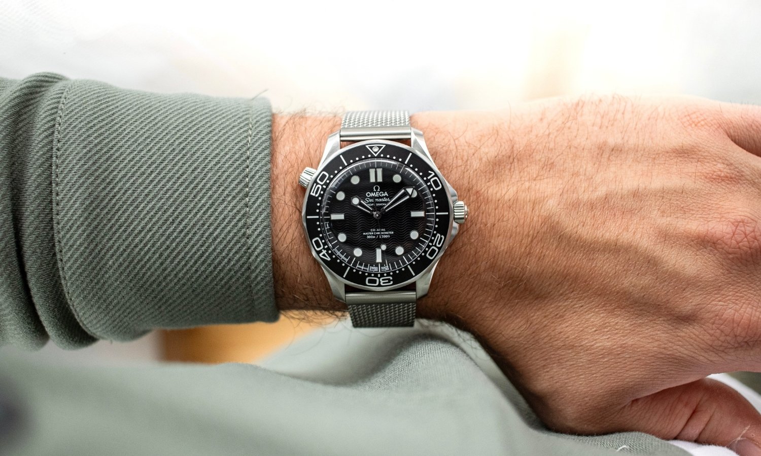 OMEGA Seamaster Diver 300M No-Date mit schwarzem Zifferblatt am Milanaise-Armband an einem Handgelenk mit grünem Ärmel getragen vor hellem Hintergrund