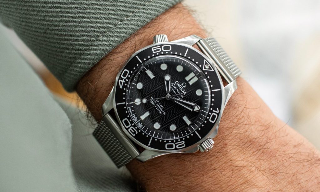 OMEGA Seamaster Diver 300M No-Date mit schwarzem Zifferblatt am Milanaise-Armband an einem Handgelenk mit grünem Ärmel getragen vor hellem Hintergrund