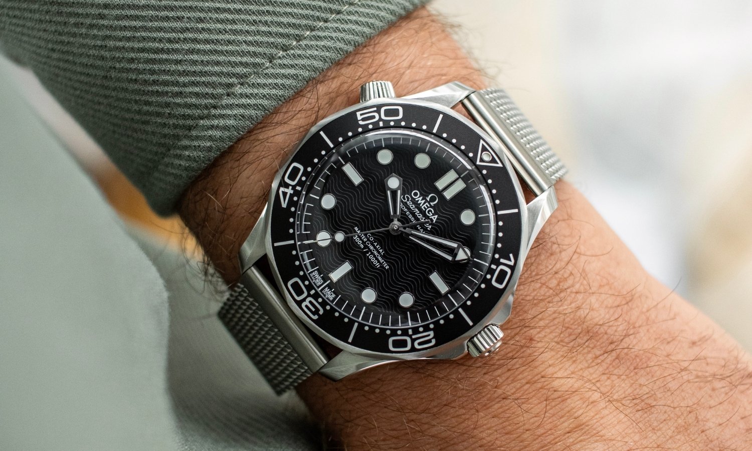 OMEGA Seamaster Diver 300M No-Date mit schwarzem Zifferblatt am Milanaise-Armband an einem Handgelenk mit grünem Ärmel getragen vor hellem Hintergrund