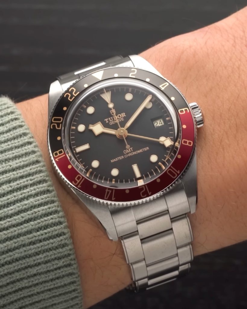 TUDOR Black Bay 58 GMT mit schwarzem Zifferblatt am Edelstahlarmband an einem Handgelenk mit grünem Ärmel getragen vor schwarzem Hintergrund