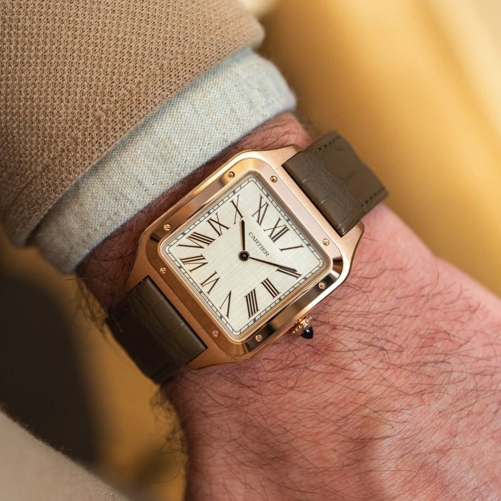 Cartier Santos Dumont in Roségold mit beigem Zifferblatt und grauem Lederarmband abgebildet an einem Handgelenk mit beigem Ärmel vor goldenem Hintergrund