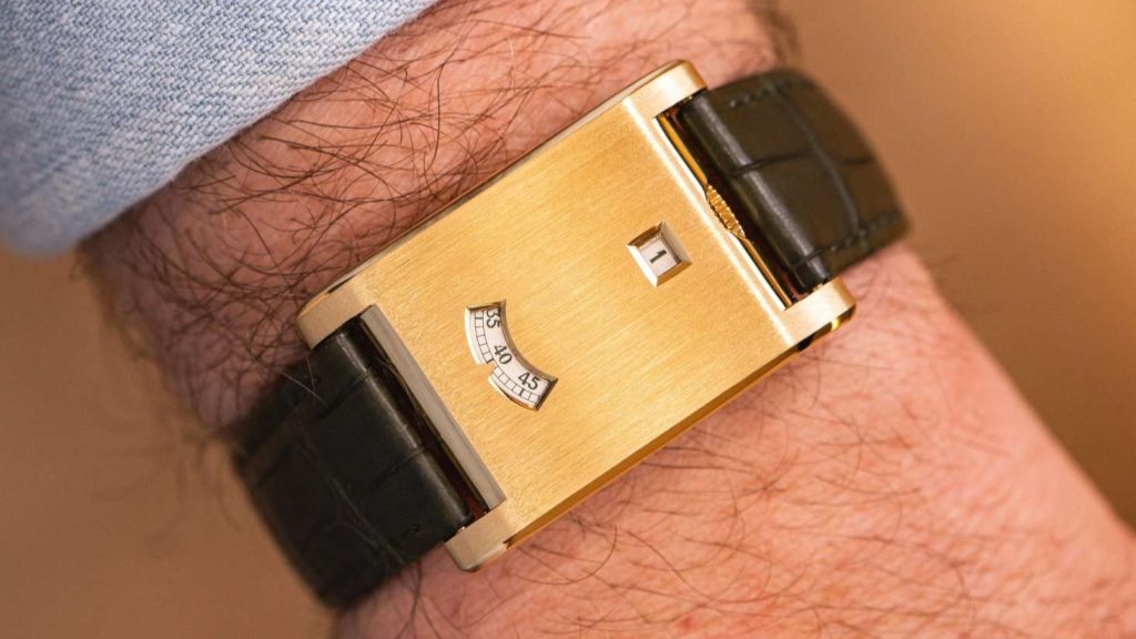 Cartier Cartier Tank a Guichets in Gelbgold mit dunkelgrünem Lederarmband abgebildet an einem Handgelenk mit hellblauem Ärmel vor goldenem Hintergrund