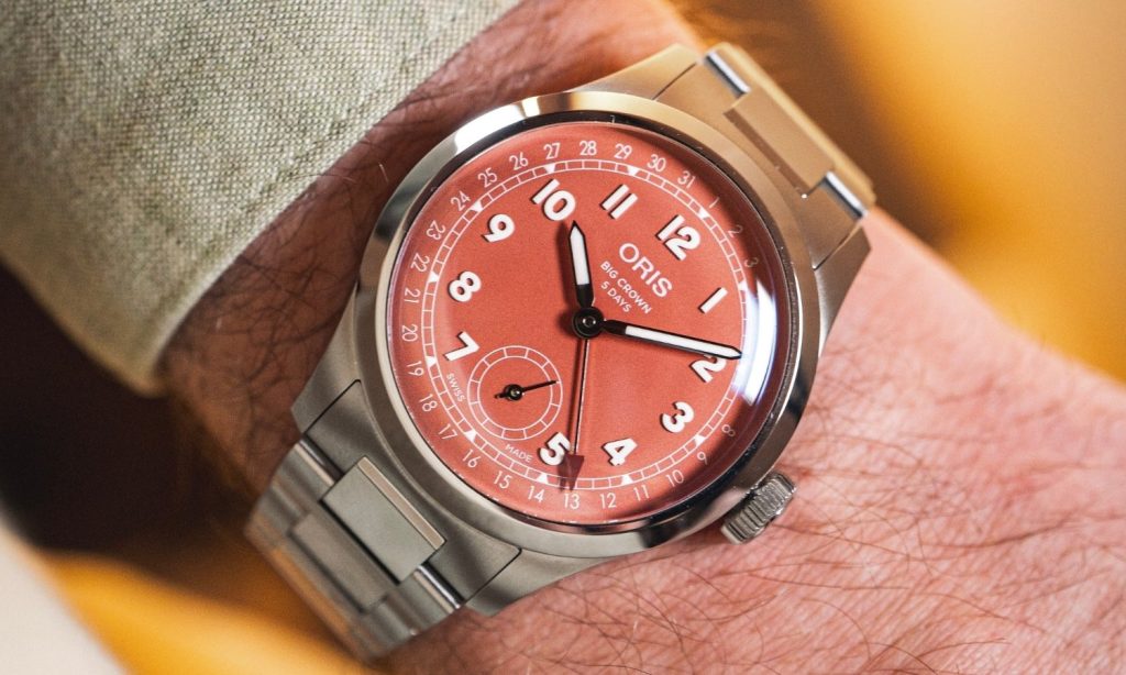 Oris Big-Crown Pointer Date mit rotem Zifferblatt am Edelstahlarmband an einem Handgelenk mit grünem Ärmel getragen vor goldenem Hintergrund
