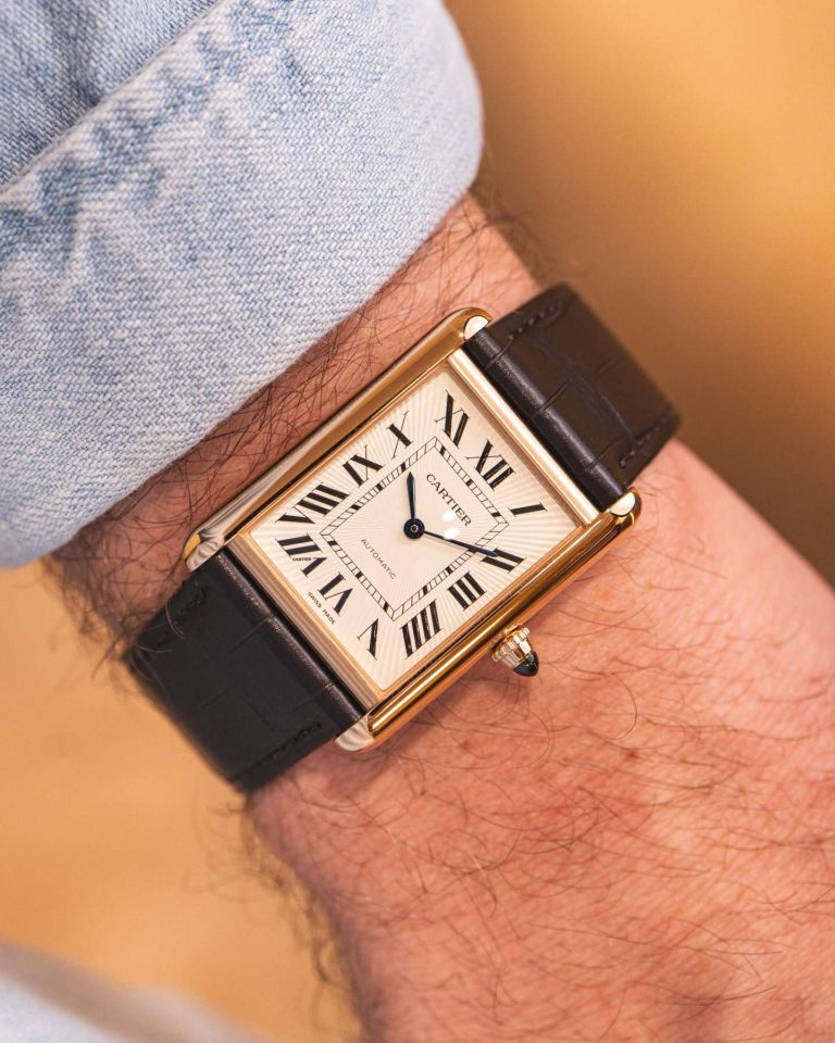 Cartier Tank Louis Cartier in Gelbgold mit dunkelgrauem Lederarmband abgebildet an einem Handgelenk mit hellblauem Ärmel vor goldenem Hintergrund