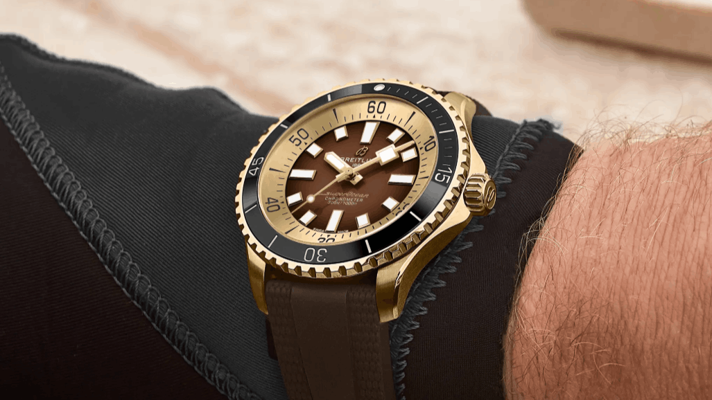 Breitling Superocean Automatic 44 in Bronze mit braunem Zifferblatt, schwarzer Lünette und braunem Kautschukband abgebildet an einem Handgelenk mit Neoprenanzug vor beigem Hintergrund