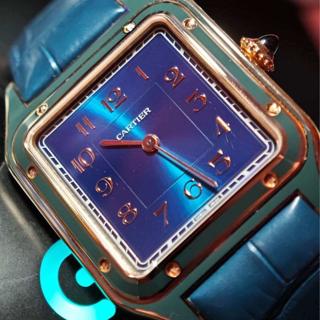 Nahaufnahme des blauen Zifferblattes der Cartier Santos-Dumont WGSA0098, die auf eine Computer-Maus der Marke Logitech gelehnt ist.