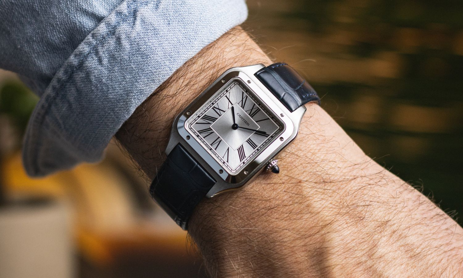 Cartier Santos-Dumont mit silbernem Zifferblatt und blauem Lederarmband abgebildet an einem Handgelenk mit hellblauem Ärmel vor dunklem Hintergrund
