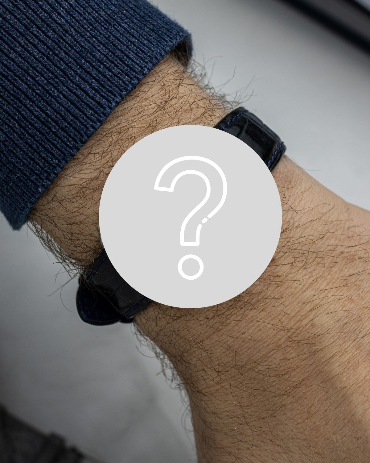 Eine Armbanduhr, die durch ein graues Fragezeichen verdeckt wird, abgebildet an einem schwarzen Lederband und einem Handgelenk mit dunkelblauem Ärmel vor weißem Hintergrund