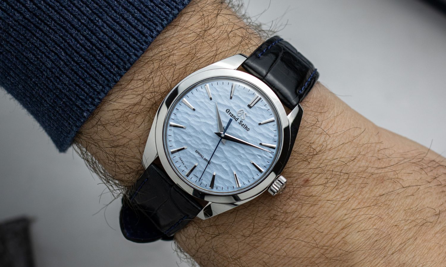 Grand Seiko Omiwatari mit blauem Zifferblatt und schwarzem Lederarmband abgebildet an einem Handgelenk mit dunklem Ärmel vor weißem Hintergrund