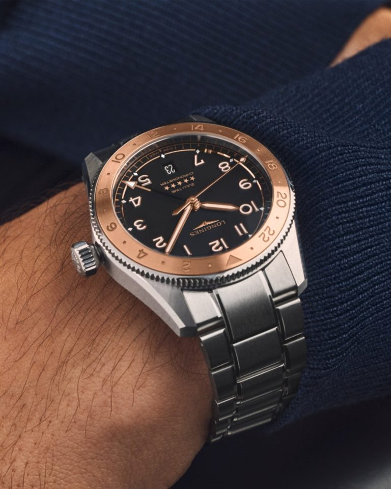 Longines Spirit Zulu Time Bicolor mit schwarzem Zifferblatt am Edelstahlarmband an einem Handgelenk mit blauem Ärmel getragen vor dunklem Hintergrund
