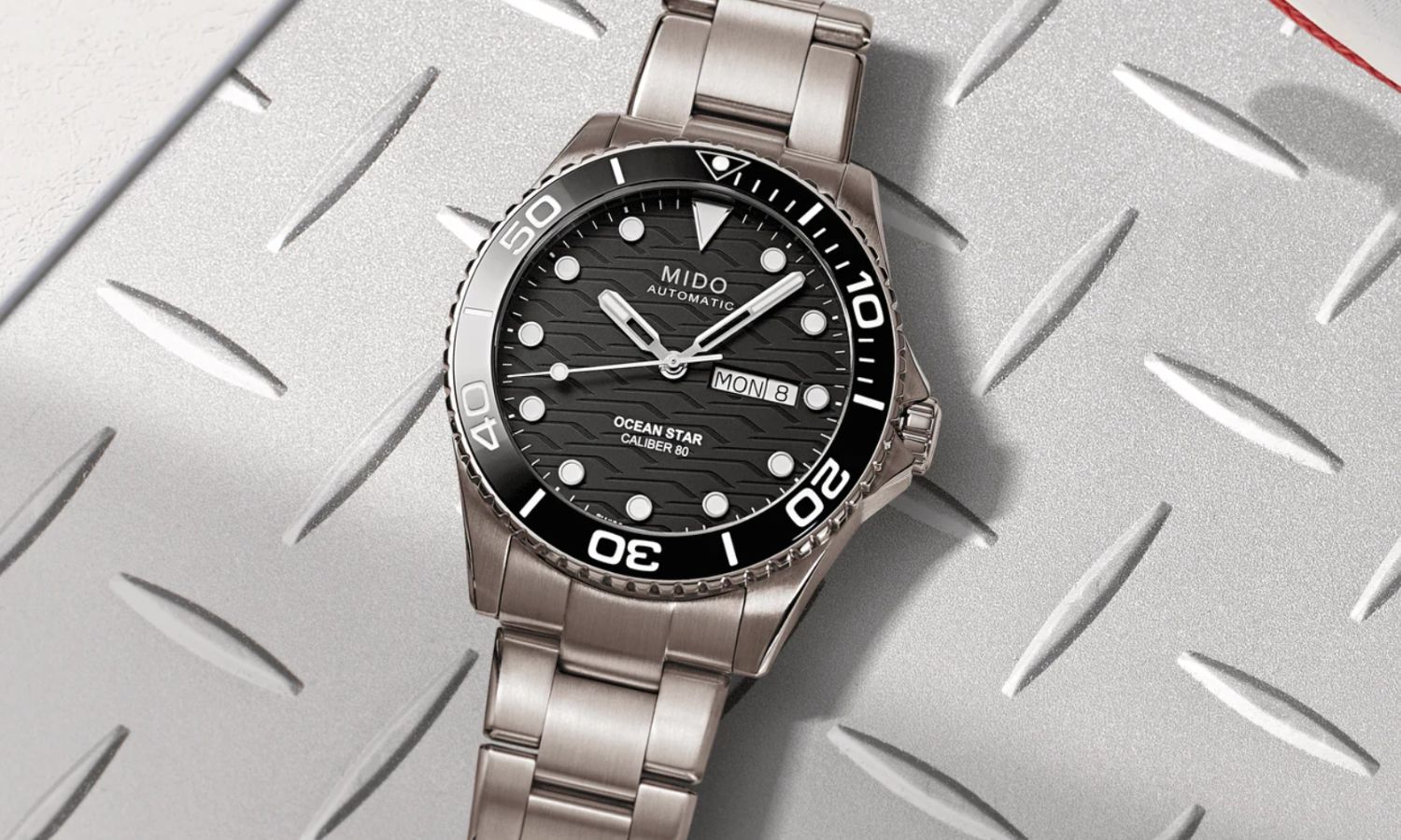 Mido Ocean Star 200 C Titan mit schwarzem Zifferblatt und schwarzer Lünette sowie einem Titanarmband abgebildet vor silbernem Hintergrund mit Struktur