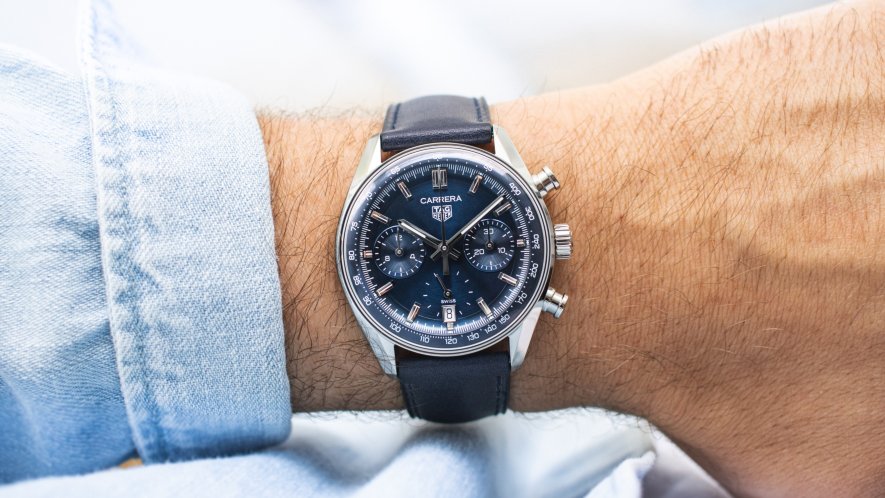 TAG Heuer Carrera Glassbox Chronograph mit blauem Zifferblatt und blauem Armband abgebildet an einem Handgelenk mit hellblauem Ärmel vor weißem Hintergrund
