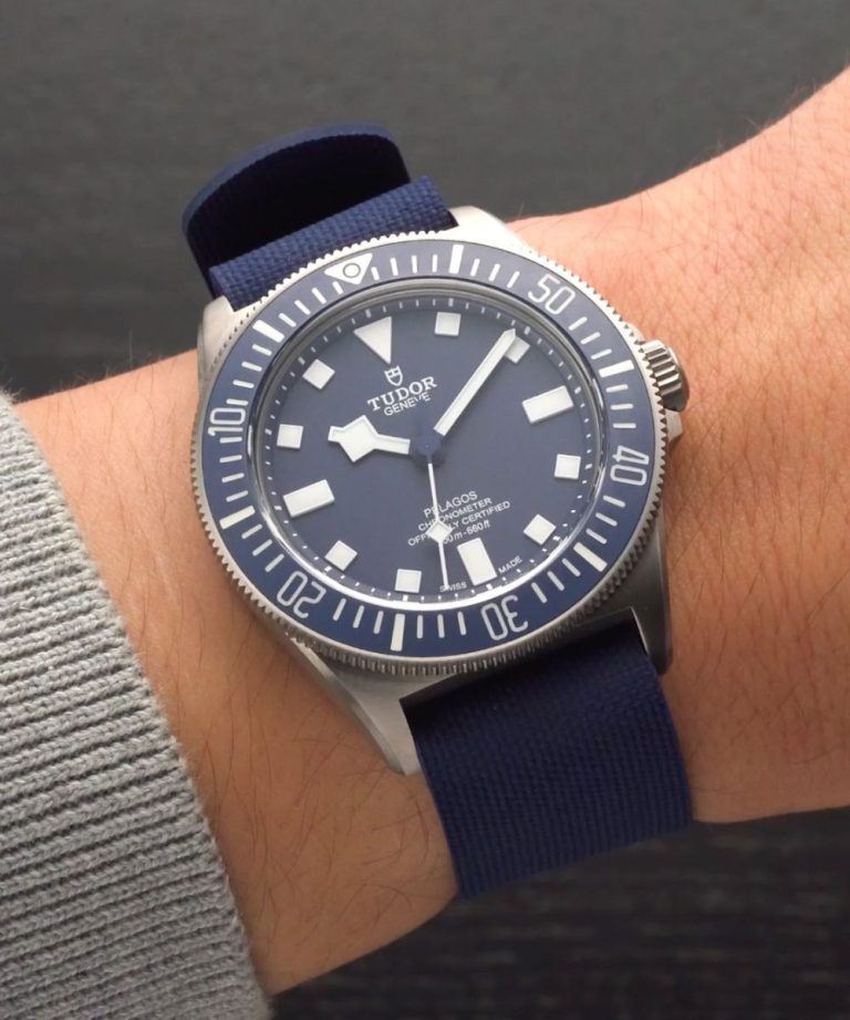 TUDOR Pelagos FXD CPO mit blauem Zifferblatt am blauen Textilarmband an einem Handgelenk mit grauem Ärmel getragen vor dunkelgrauem Hintergrund