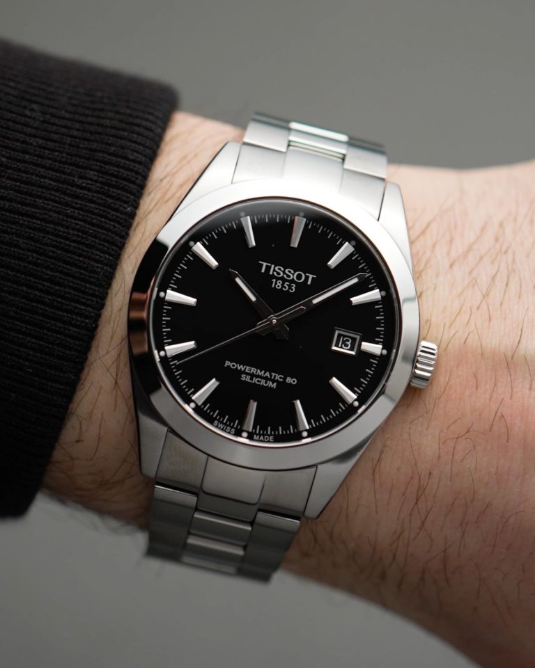 Tissot Gentleman Powermatic 80 Referenz: T127.407.11.051.00 mit schwarzem Zifferblatt am Edelstahlarmband an einem Handgelenk mit schwarzem Ärmel getragen vor grauem Hintergrund