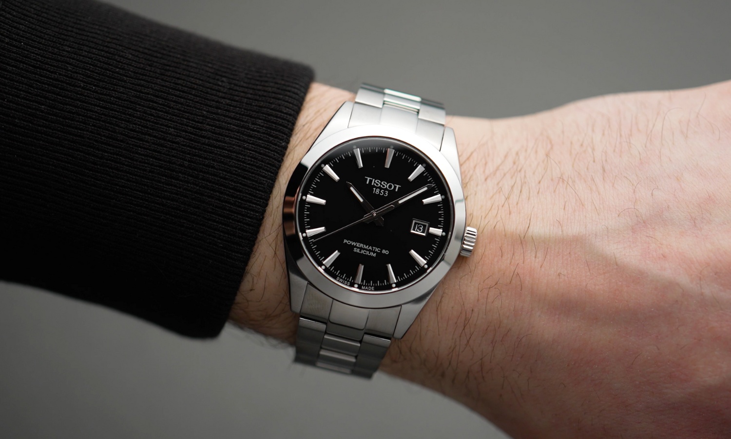 Tissot Gentleman Powermatic 80 Referenz: T127.407.11.051.00 mit schwarzem Zifferblatt am Edelstahlarmband an einem Handgelenk mit schwarzem Ärmel getragen vor grauem Hintergrund