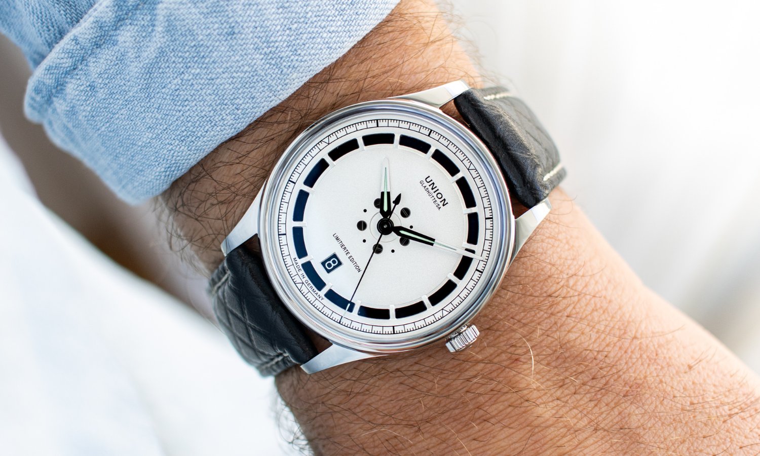 UNION Glashütte Noramis Datum Limitierte Edition Deutschland Klassik 2025 mit silbernem Zifferblatt am schwarzen Lederarmband an einem Handgelenk mit hellblauem Ärmel getragen vor hellem Hintergrund