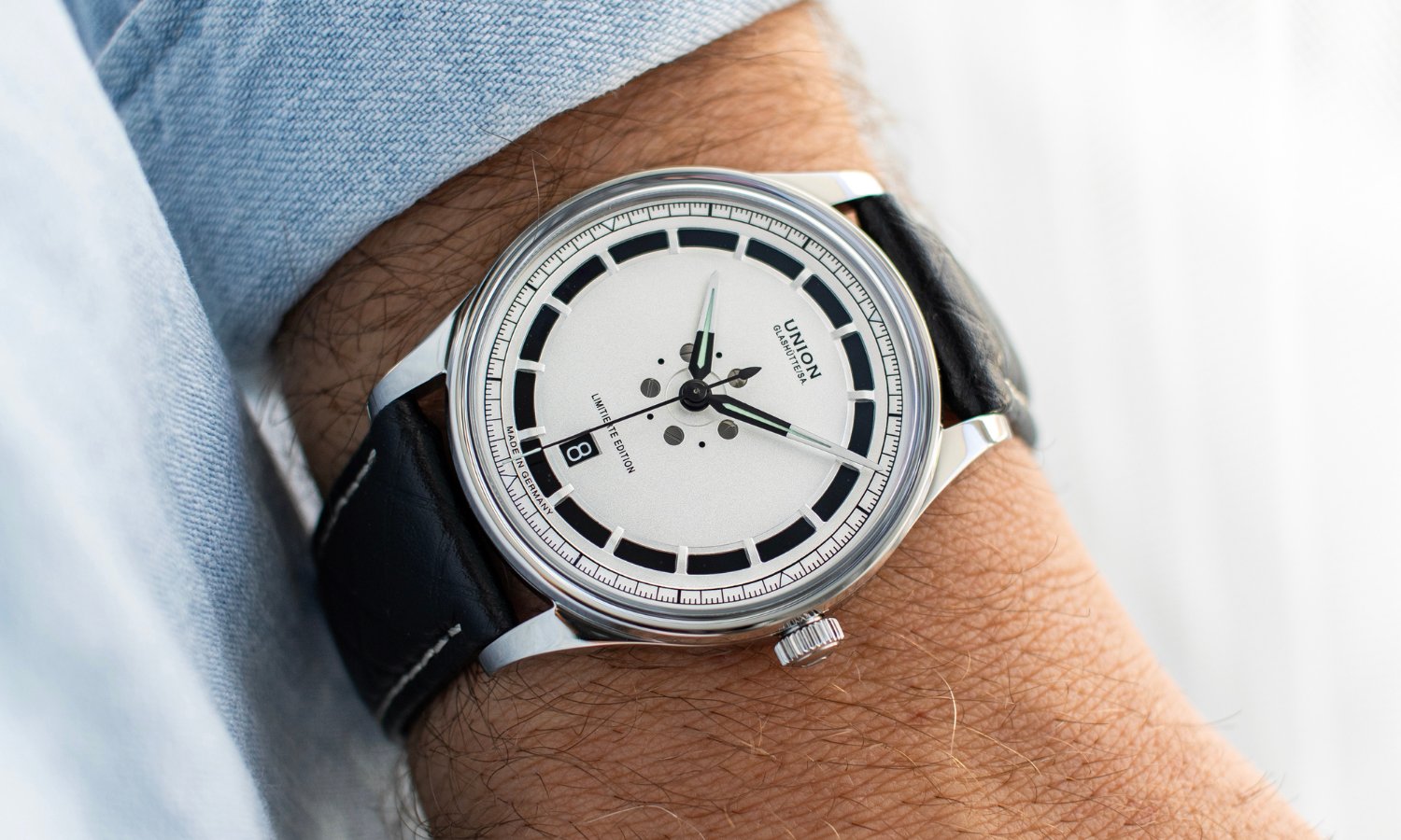 UNION Glashütte Noramis Datum Limitierte Edition Deutschland Klassik 2025 mit silbernem Zifferblatt am schwarzen Lederarmband an einem Handgelenk mit hellblauem Ärmel getragen vor hellem Hintergrund