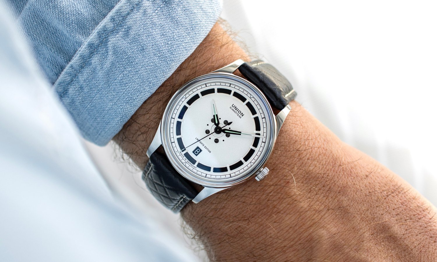UNION Glashütte Noramis Datum Limitierte Edition Deutschland Klassik 2025 mit silbernem Zifferblatt am schwarzen Lederarmband an einem Handgelenk mit hellblauem Ärmel getragen vor hellem Hintergrund