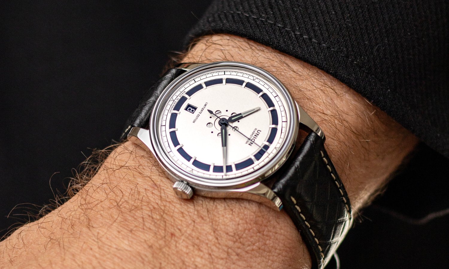 UNION Glashütte Noramis Datum Limitierte Edition Deutschland Klassik 2025 mit silbernem Zifferblatt am schwarzen Lederarmband an einem Handgelenk mit schwarzem Ärmel getragen vor schwarzem Hintergrund