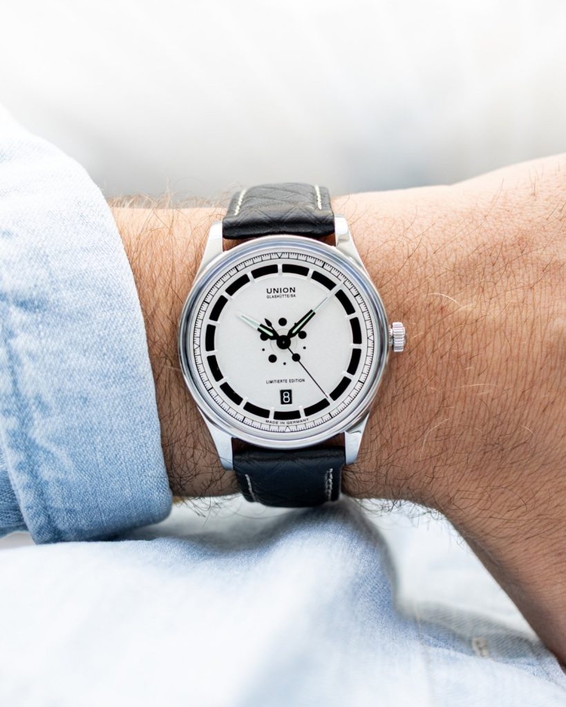 UNION Glashütte Noramis Datum Limitierte Edition Deutschland Klassik 2025 mit silbernem Zifferblatt am schwarzen Lederarmband an einem Handgelenk mit hellblauem Ärmel getragen vor hellem Hintergrund