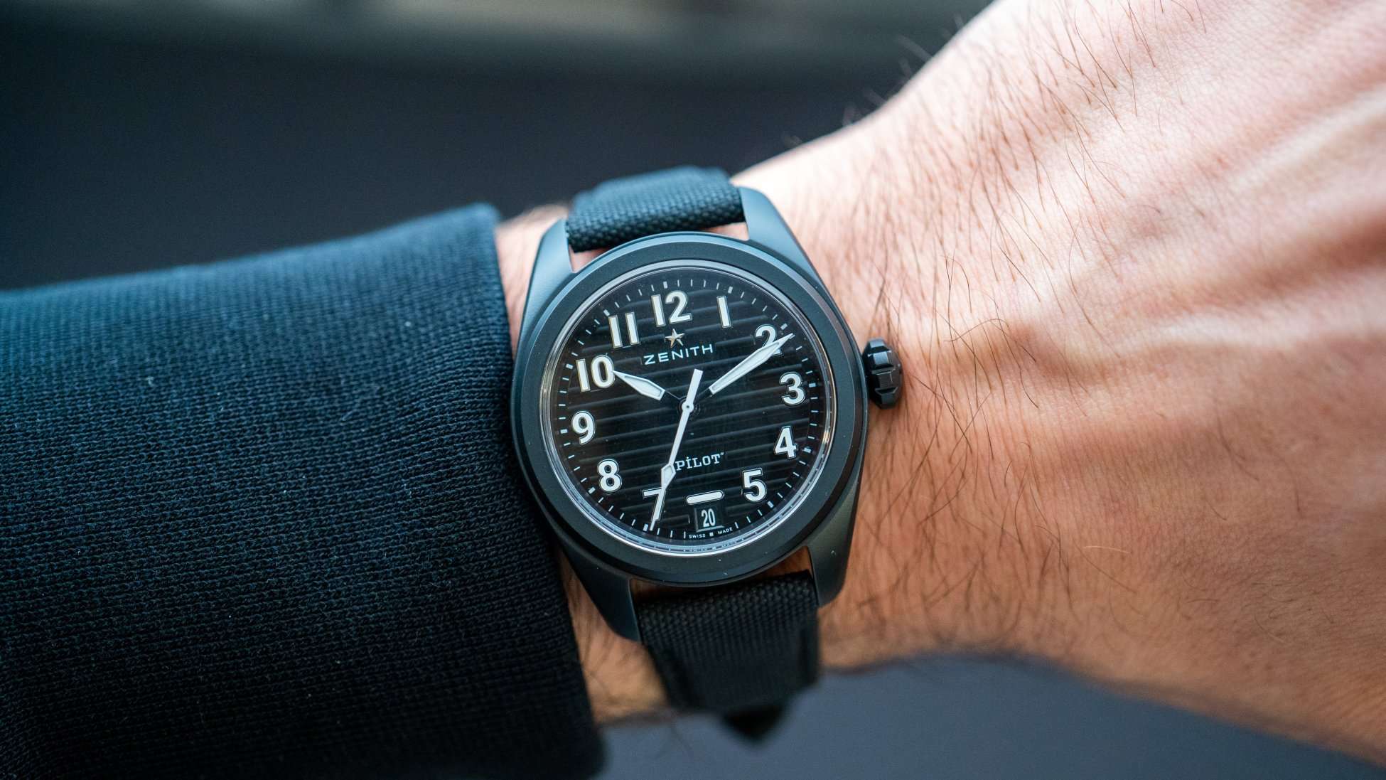 Zenith Pilot Automatik mit schwarzem Keramikgehäuse, schwarzem Zifferblatt und schwarzem Textilarmband abgebildet an einem Handgelenk mit schwarzem Ärmel vor schwarzem Hintergrund