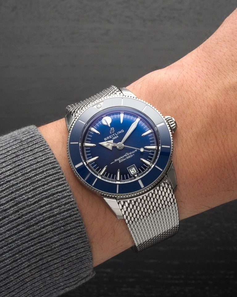 Breitling SuperOcean Heritage B31 mit blauem Zifferblatt am Milanaise Armband an einem Handgelenk mit grauem Ärmel getragen vor grauem Hintergrund