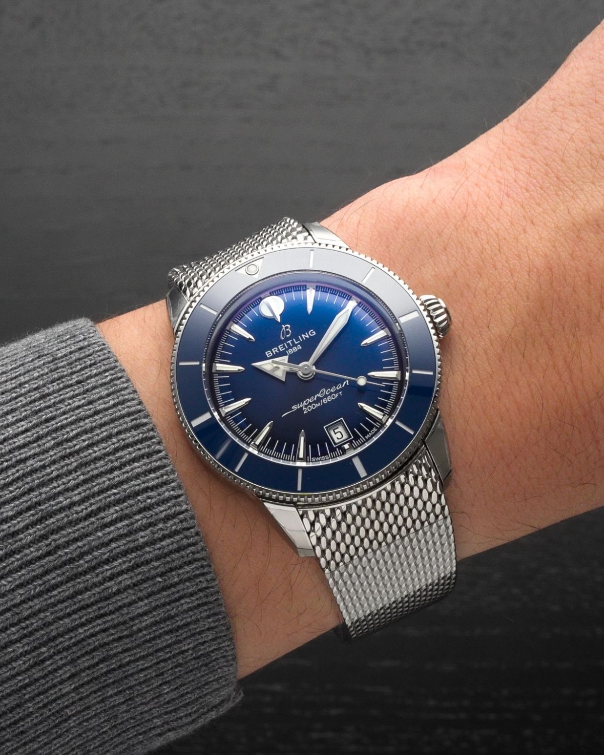 Breitling SuperOcean Heritage B31 mit blauem Zifferblatt am Milanaise Armband an einem Handgelenk mit grauem Ärmel getragen vor grauem Hintergrund