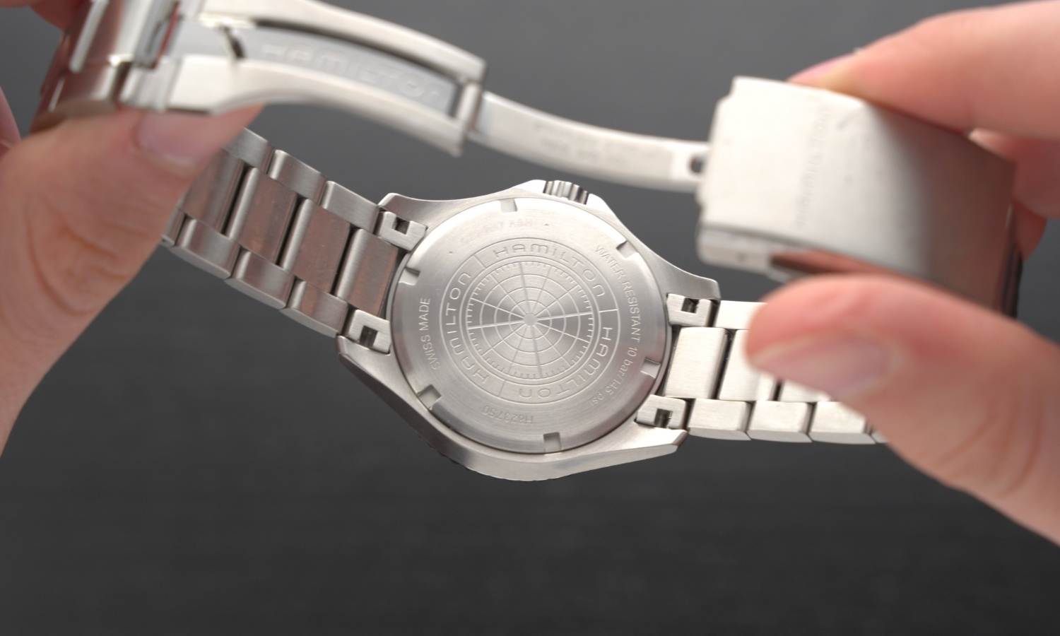 Der Gehäuseboden und das Edelstahlarmband der Hamilton Khaki Navy Scuba Auto Referenz: H82375161 in zwei Händen gehalten vor dunkelgrauem Hintergrund