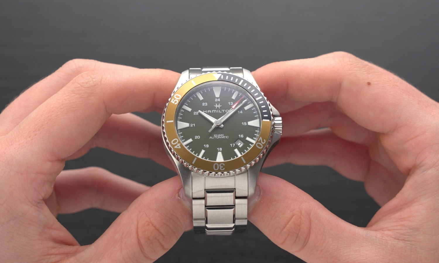Hamilton Khaki Navy Scuba Auto Referenz: H82375161 mit grünem Zifferblatt am Edelstahlarmband in zwei Händen gehalten vor dunkelgrauem Hintergrund