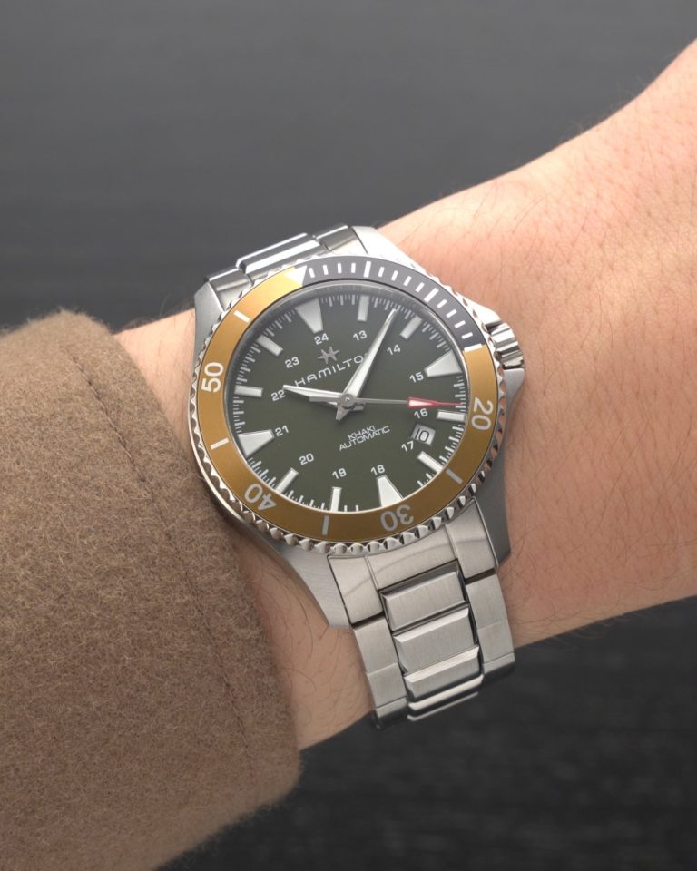 Hamilton Khaki Navy Scuba Auto Referenz: H82375161 mit grünem Zifferblatt am Edelstahlarmband an einem Handgelenk mit braunem Ärmel getragen vor dunkelgrauem Hintergrund