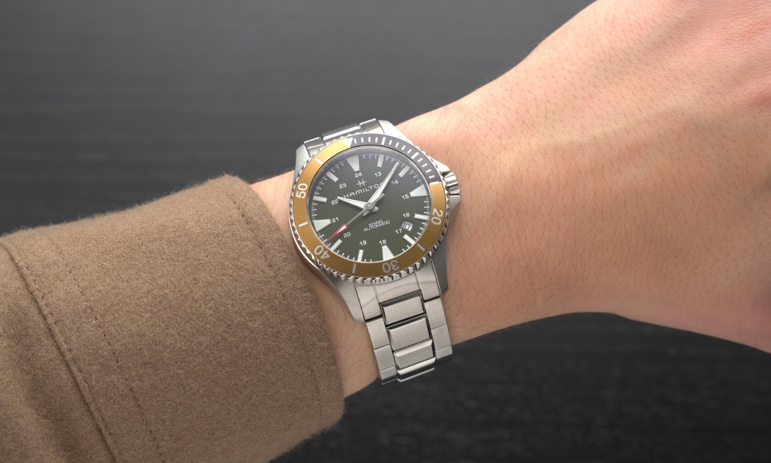 Hamilton Khaki Navy Scuba Auto Referenz: H82375161 mit grünem Zifferblatt am Edelstahlarmband an einem Handgelenk mit braunem Ärmel getragen vor dunkelgrauem Hintergrund