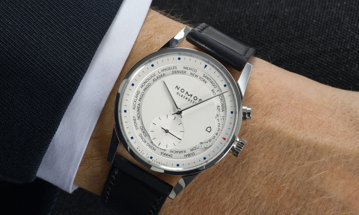 NOMOS Glashütte Zürich Weltzeit mit weißem Zifferblatt und schwarzem Lederarmband abgebildet an einem Handgelenk mit dunklem Jackett und weißem Hemd vor dunklem Hintergrund